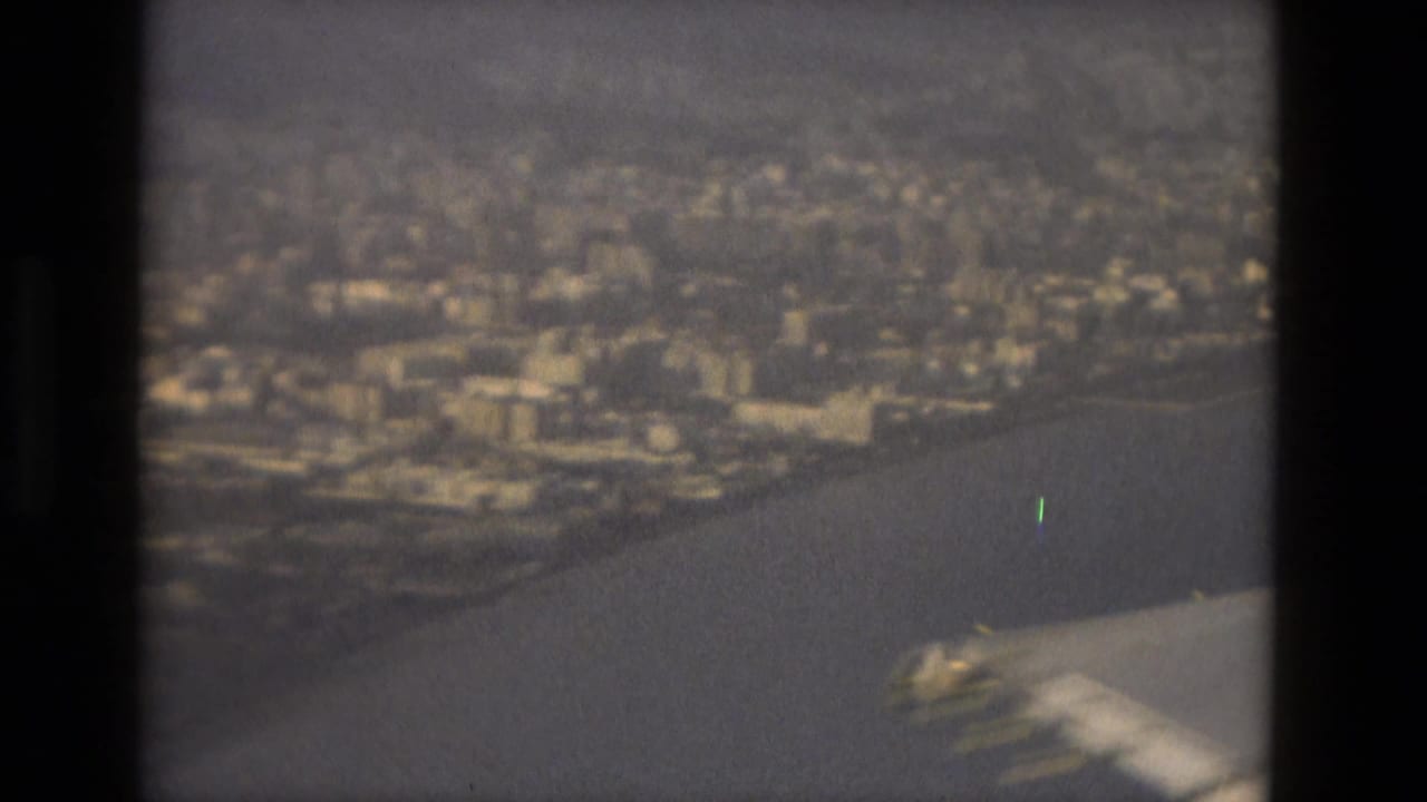 1978 8mm Barcelona travel footage appears to show an airplane wing above coastline, city blocks, and water in soft haze.