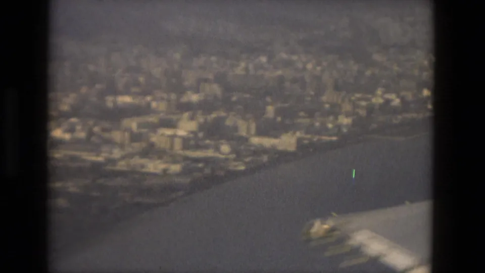 1978 8mm Barcelona travel footage appears to show an airplane wing above coastline, city blocks, and water in soft haze.
