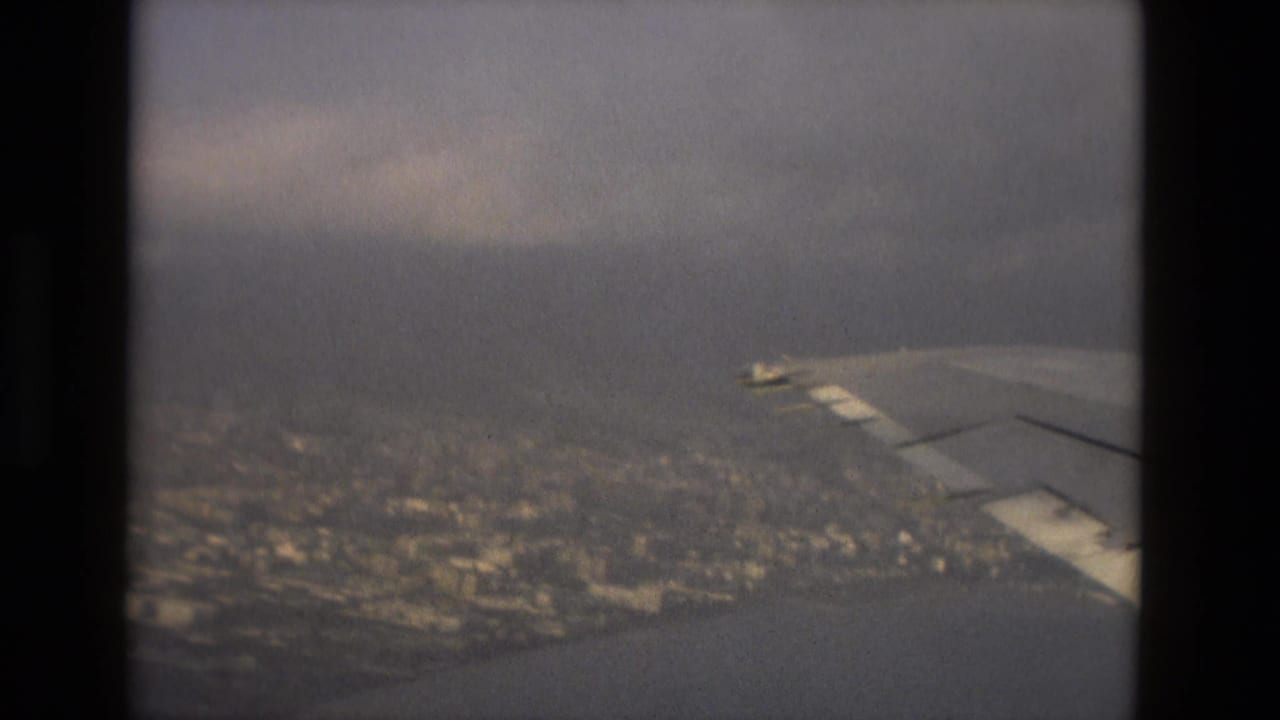 A passenger view from an aircraft shows the wing over Barcelona's coastal urban area, water, and low cloud haze.