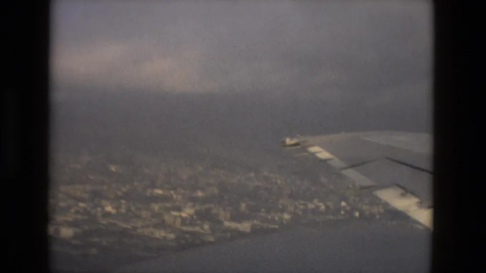 A passenger view from an aircraft shows the wing over Barcelona's coastal urban area, water, and low cloud haze.