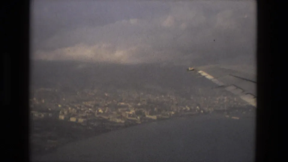 1978 8mm footage shows an aircraft wing over a hazy Barcelona coastal view with dense city blocks and water below.