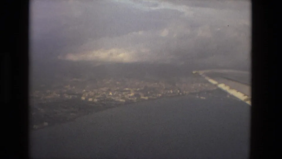 The airplane wing frames a muted 8mm aerial view of Barcelona, with water, shoreline, city texture, and heavy clouds visible.