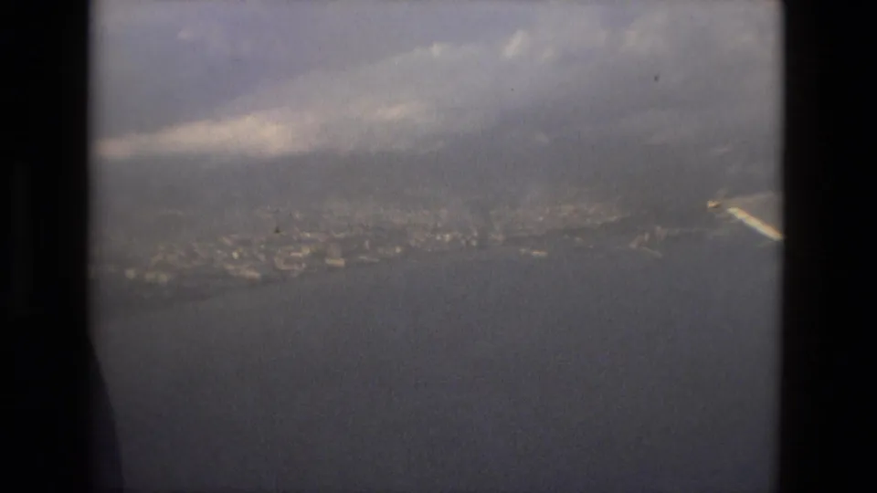 1978 8mm airplane-window view over open water toward the distant Barcelona shoreline under cloud cover.