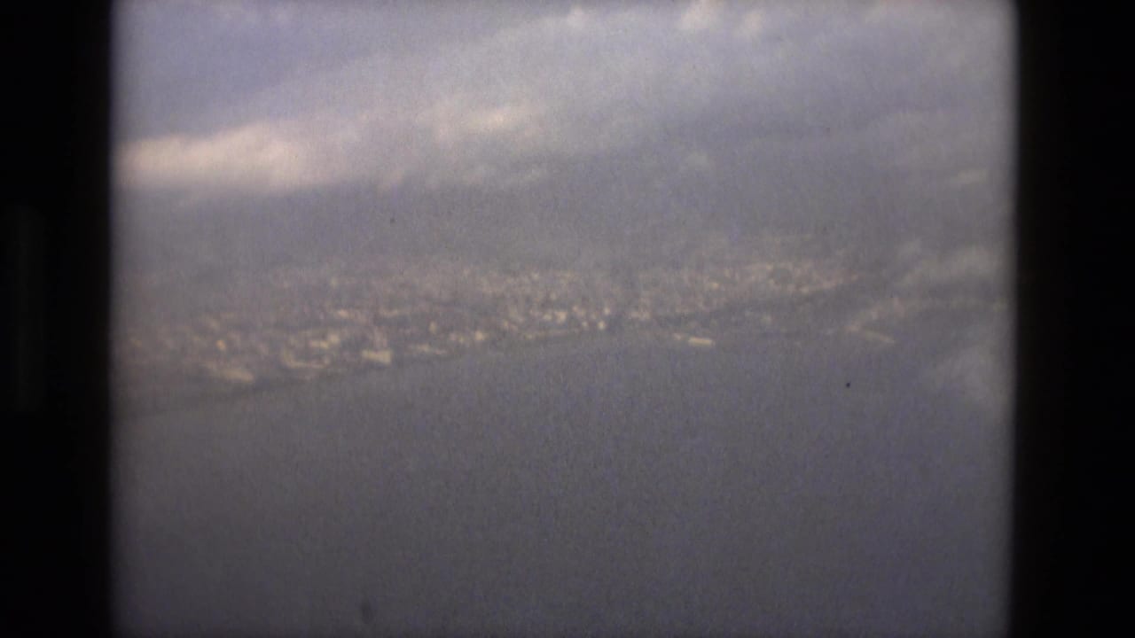 A soft 8mm view from an airplane window shows Barcelona's distant coast across water beneath a clouded sky.