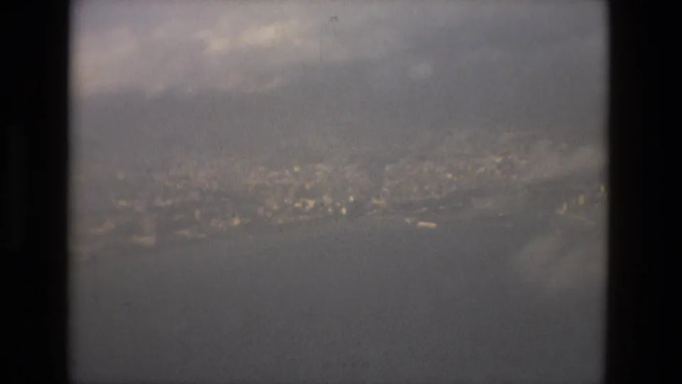 A grainy 1978 home-movie frame shows the Barcelona coast and water through the dark edges of an airplane window.
