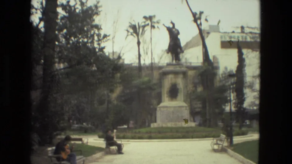 1978 Barcelona 8mm frame of a public park plaza with seated visitors, benches, palms, greenery, and an equestrian statue.