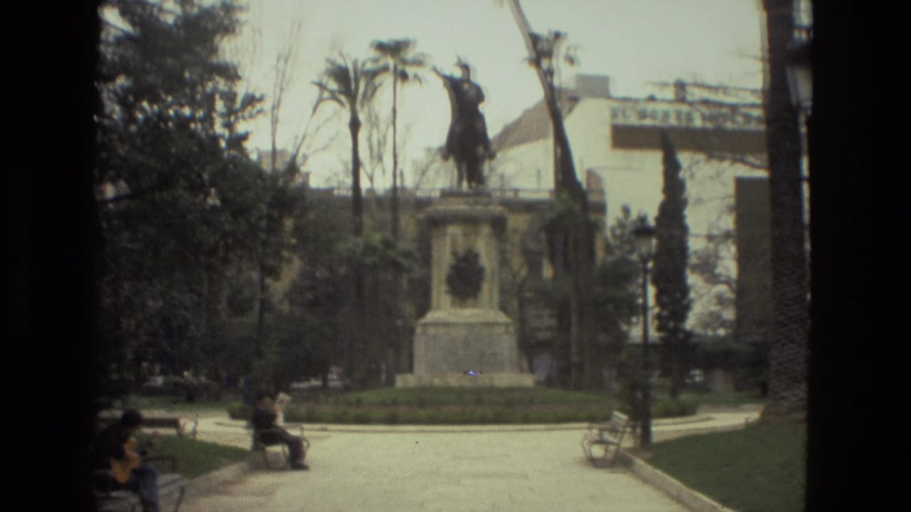 1978 Barcelona home-movie frame showing an equestrian statue on a pedestal in a park, with benches, trees, and visitors in view.