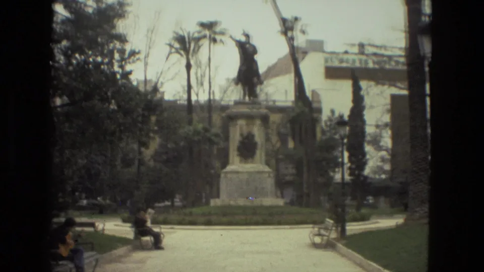 1978 Barcelona home-movie frame showing an equestrian statue on a pedestal in a park, with benches, trees, and visitors in view.