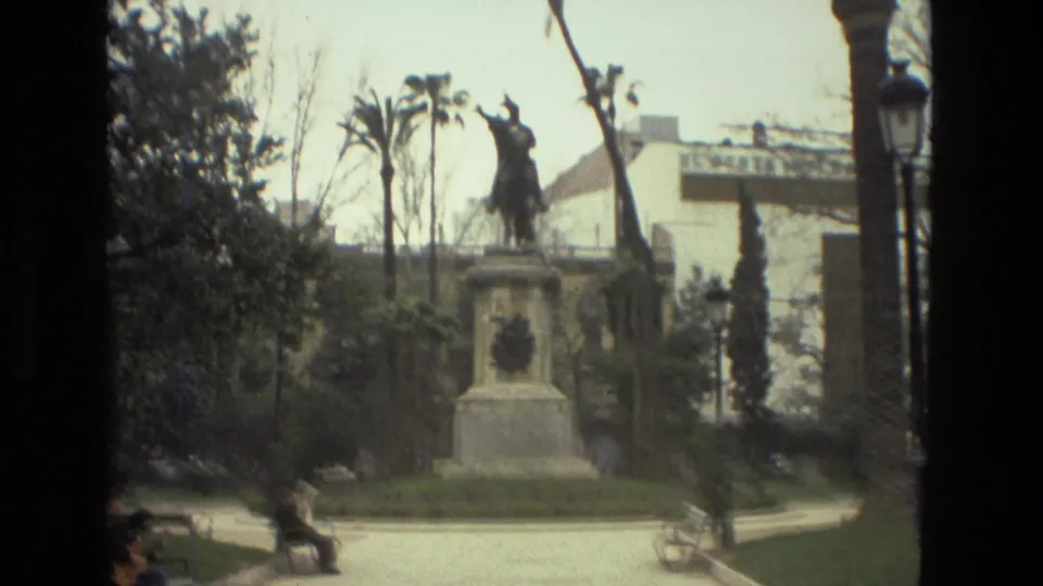 1978 Barcelona park view with a mounted statue, palm trees, a lamppost, benches, and a blurred person passing through the plaza.