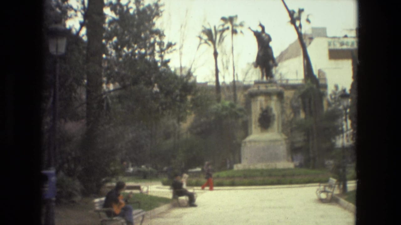 People sit on benches in a park near an equestrian statue and palm trees.
