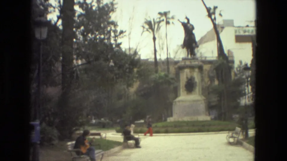 1978 Barcelona home-movie frame showing people seated in a park, with a guitarist on a bench and an equestrian statue behind them.
