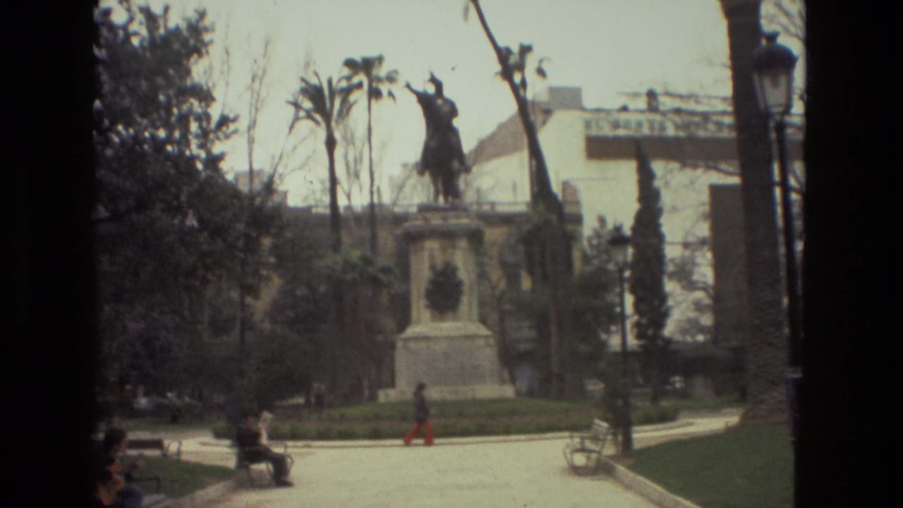 Barcelona park scene on 8mm film, with seated visitors, a person walking across the square, trees, and an equestrian statue in the background.