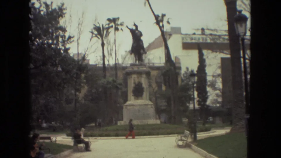 Barcelona park scene on 8mm film, with seated visitors, a person walking across the square, trees, and an equestrian statue in the background.