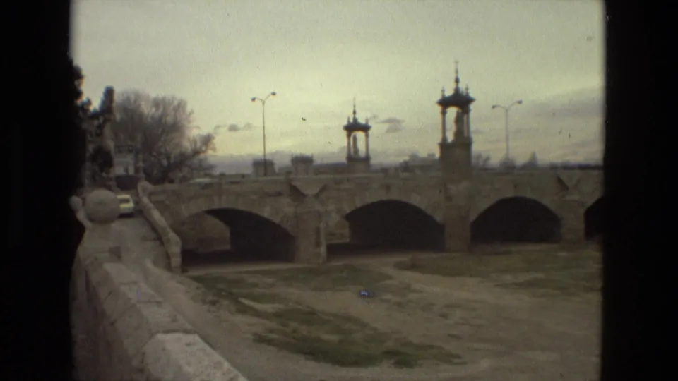 1978 Barcelona travel footage view of a stone arched bridge with small tower structures, seen from a moving roadside or walkway angle.