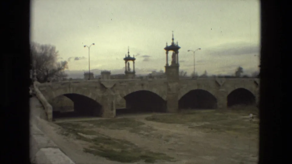 A soft 8mm frame of a historic-looking stone bridge in Barcelona, with multiple arches, small towers, and open ground below.