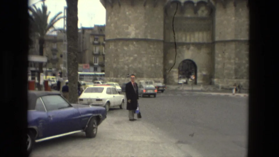 A suited man in sunglasses stands near a blue convertible and white car outside a Barcelona stone monument in 1978.