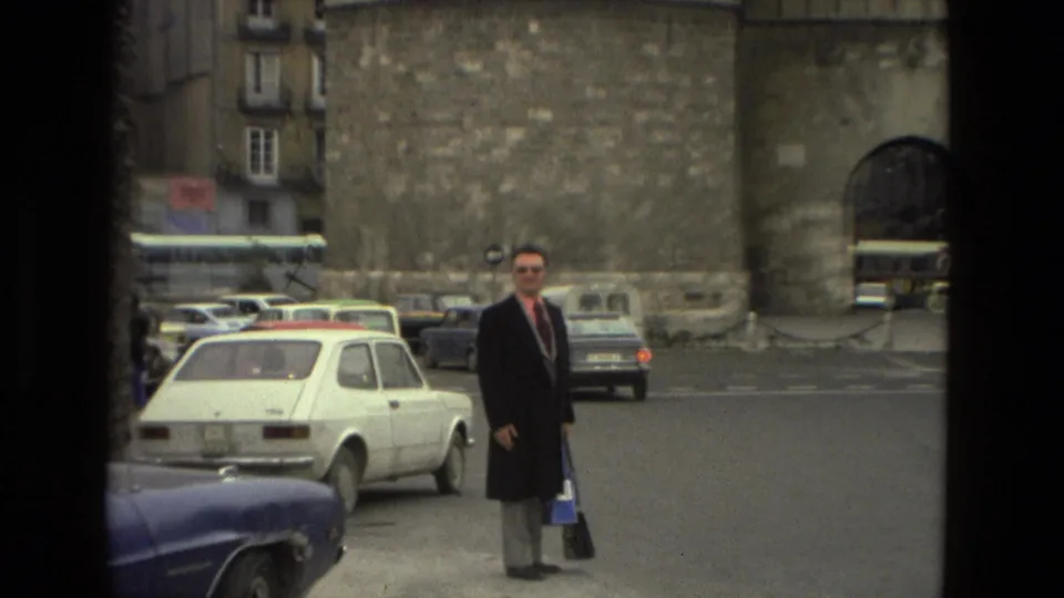 Closer 1978 Barcelona view of a suited adult man beside traffic, with stonework and an arched monument entrance behind him.