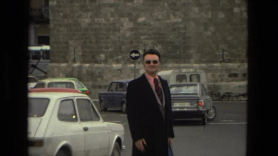 A suited man in sunglasses smiles toward the camera in a 1978 Barcelona street scene with compact cars and stone monument walls.