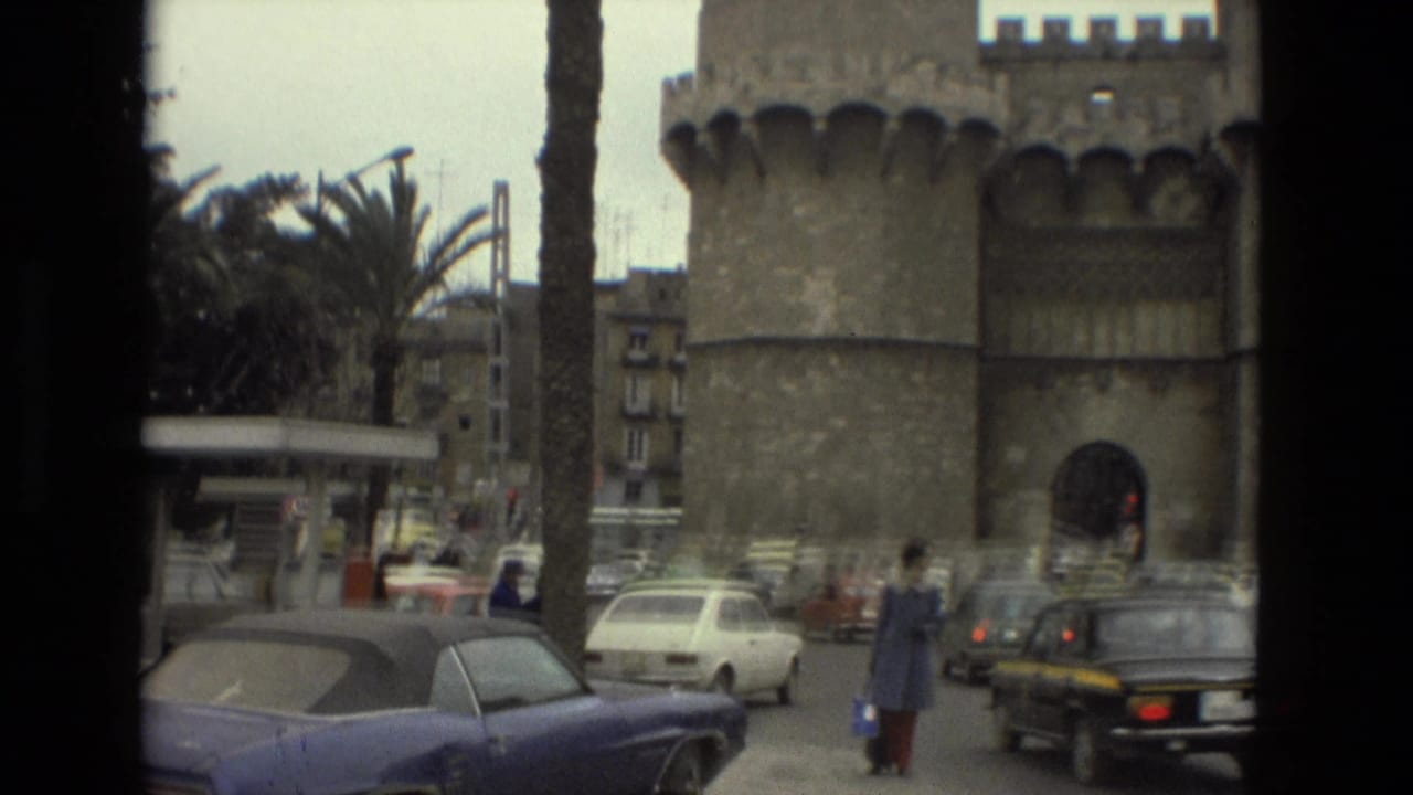 Stone gate with round towers, palm trees, cars, and a pedestrian crossing a Barcelona street.