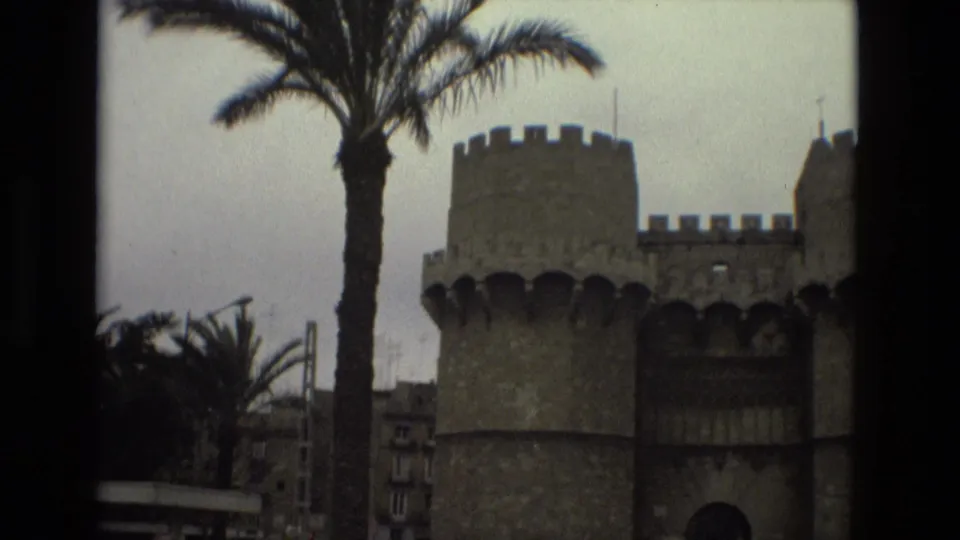 1978 Barcelona frame with a palm tree in front of a crenellated stone gate and surrounding urban buildings.