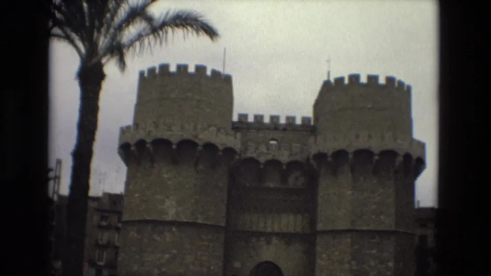 1978 Barcelona home-movie view of twin crenellated stone towers, a palm trunk, and the dark central gate opening.
