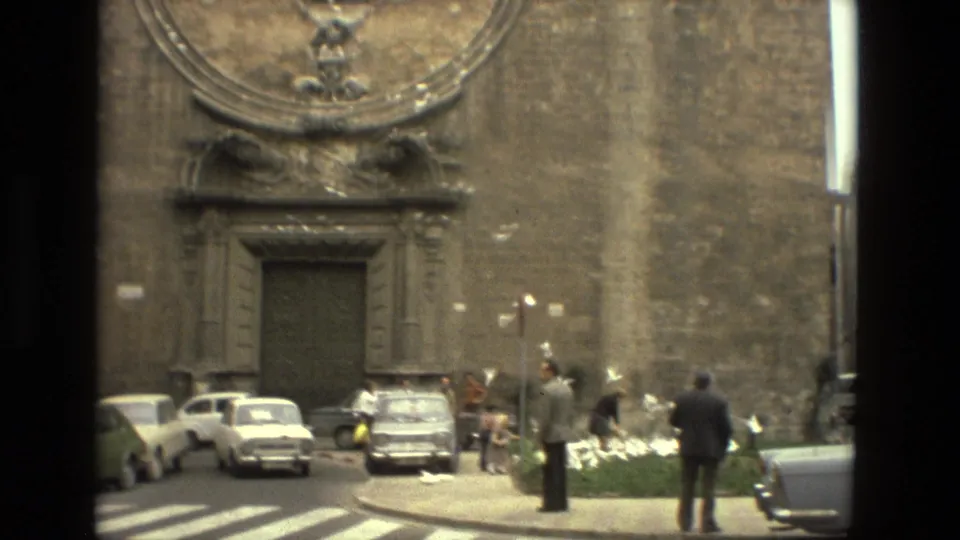 Barcelona street scene in 1978 with a stone church wall, parked 1970s cars, pedestrians, birds, and a small planted area.