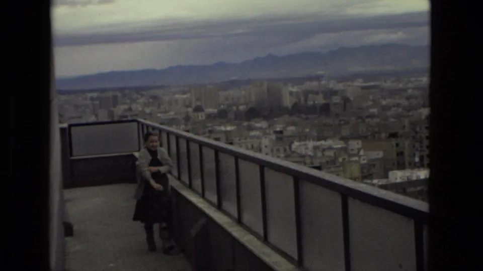 Elevated Barcelona view from a balcony, with a person near the railing and the city stretching toward distant mountains.