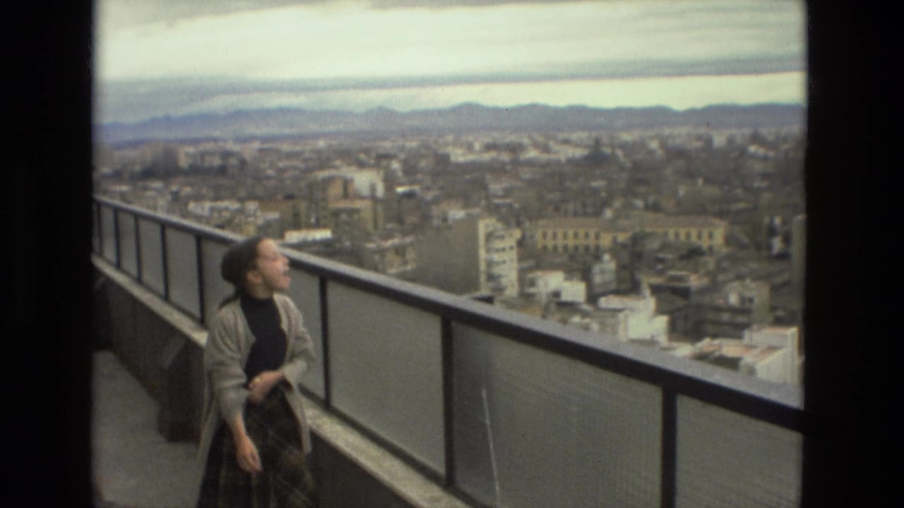 In Barcelona, Spain, a young woman in a cardigan and plaid skirt stands by a balcony railing above the city in 1978 home-movie footage.