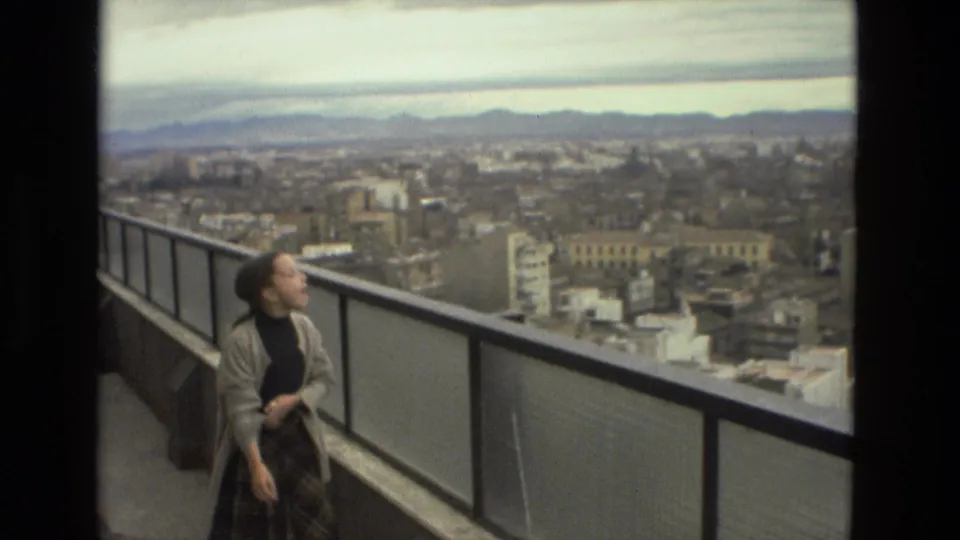 In Barcelona, Spain, a young woman in a cardigan and plaid skirt stands by a balcony railing above the city in 1978 home-movie footage.