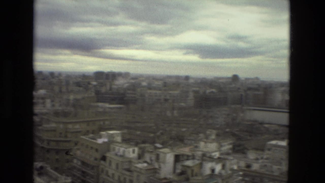 1978 Barcelona cityscape seen from a high window, with cloudy sky, rooftops, and dense mid-rise buildings in soft 8mm focus.