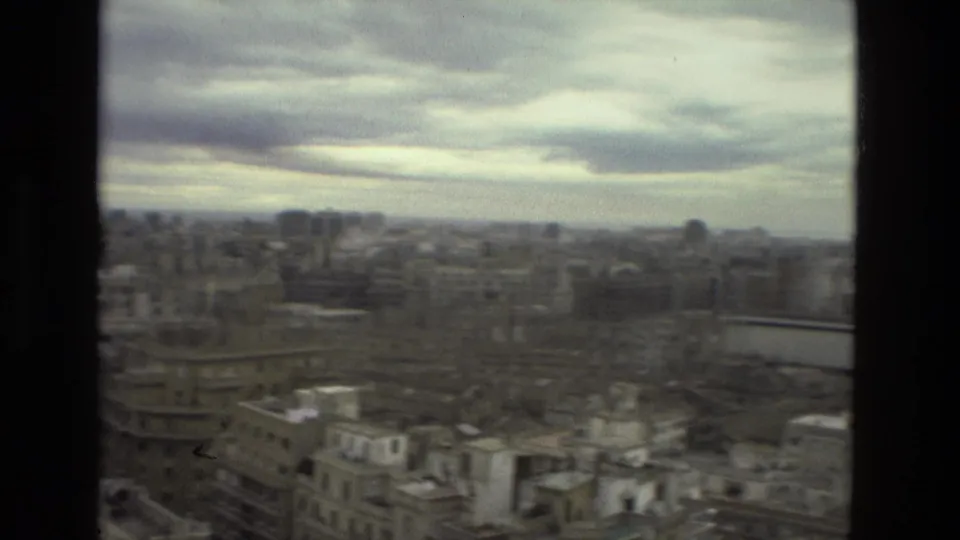 1978 Barcelona cityscape seen from a high window, with cloudy sky, rooftops, and dense mid-rise buildings in soft 8mm focus.