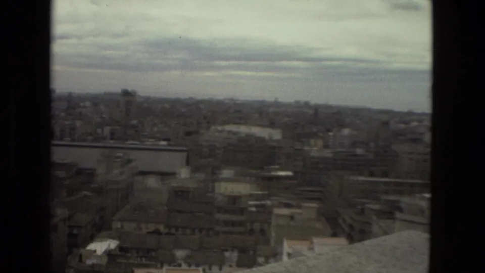 Barcelona skyline seen from above in 1978, with compact urban blocks, a pale horizon, and soft 8mm focus through a window-like frame.