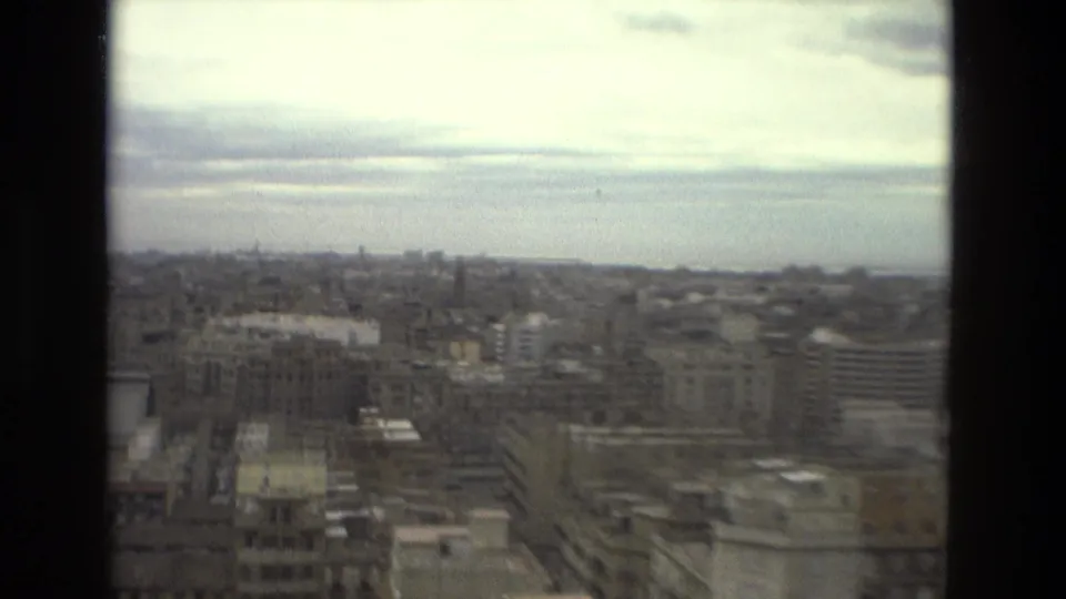 High-angle 1978 Barcelona view showing city blocks, rooftops, distant structures, and a bright overcast sky in grainy 8mm color.