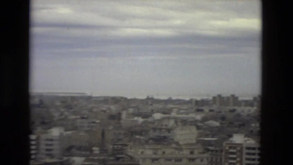 End of the 1978 Barcelona pan, with rooftops, low city structures, gray cloud cover, and a pale distant horizon in faded 8mm color.