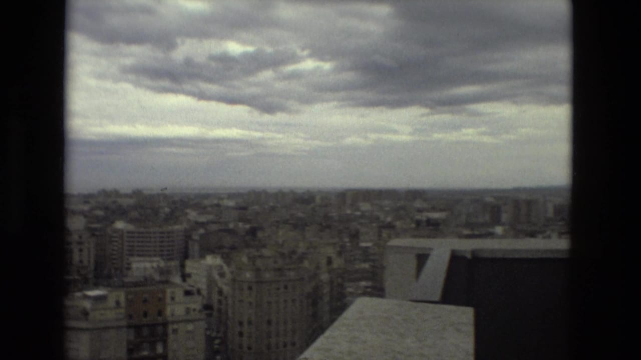 High view over Barcelona buildings beneath an overcast sky, partly framed by a dark window edge.