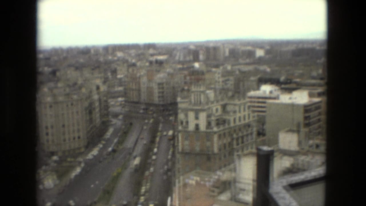 1978 Barcelona home-movie frame looking from a high-rise over a broad street, apartment blocks, rooftops, and distant urban horizon.