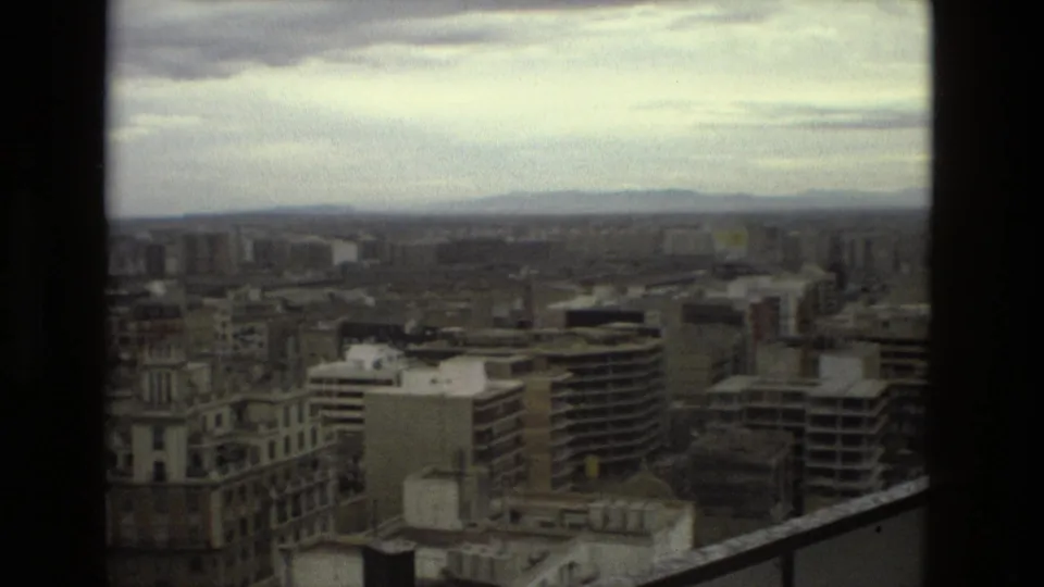 Elevated 1978 Barcelona view across mid-rise apartment blocks and rooftops toward distant hills beneath a clouded sky.
