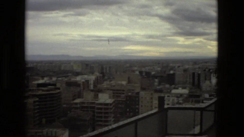 1978 Barcelona panoramic frame showing apartment towers, rooftops, cloudy horizon, and a high-rise balcony railing in the foreground.