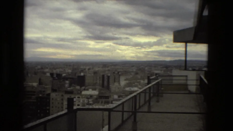 High-rise balcony view over 1978 Barcelona, with railing, building edge, dense city blocks, distant hills, and dramatic cloud cover.