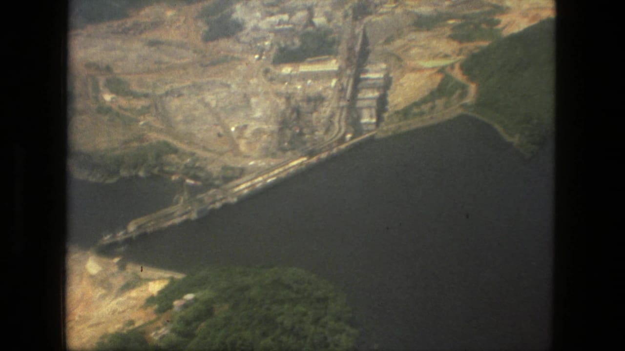 1979 Manaus, Brazil aerial frame with a large riverside industrial site, pale earthworks, dark water, and surrounding green forest.