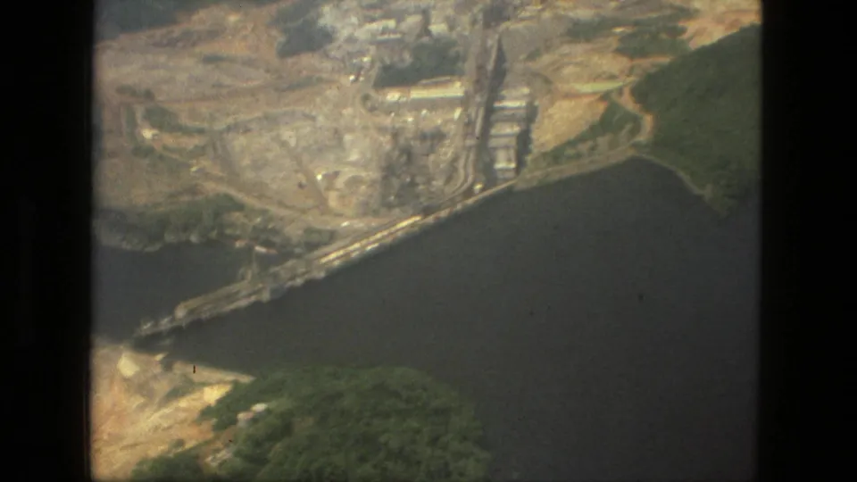1979 Manaus, Brazil aerial frame with a large riverside industrial site, pale earthworks, dark water, and surrounding green forest.