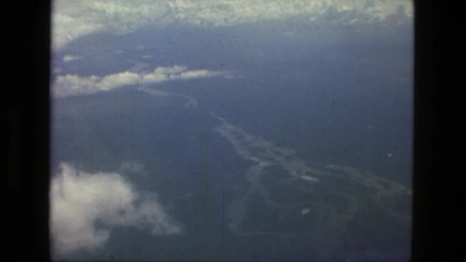 1979 Manaus, Brazil aerial 8mm frame showing broad blue water, forested shoreline, haze, and clouds from a high aircraft angle.