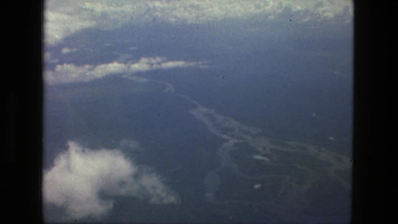 1979 8mm aerial view near Manaus, Brazil, looking past aircraft window edges toward clouds, water, and forested terrain below.