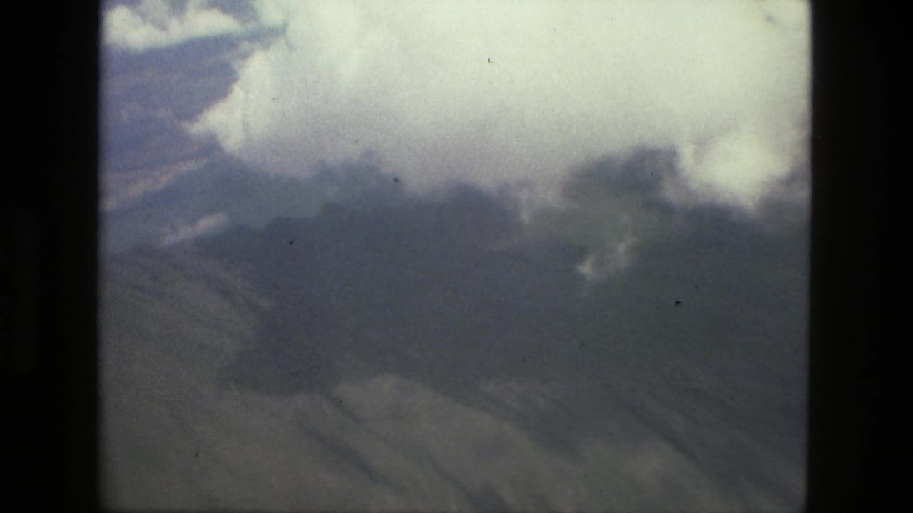 Clouds over hazy forested mountains seen from an aircraft window.