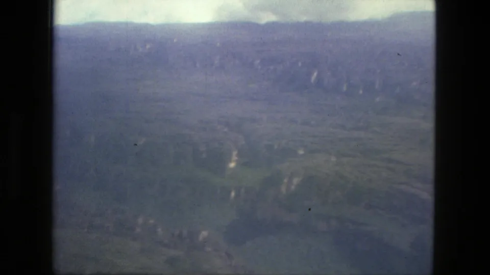 Aerial 8mm frame from the Manaus, Brazil reel with hazy ridgelines, green terrain, and pale winding breaks in the landscape.