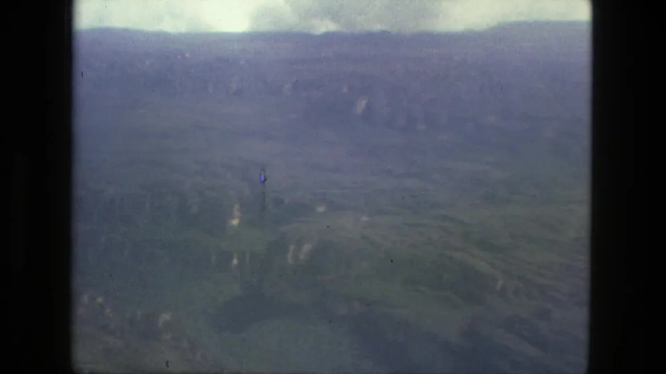 1979 home-movie aerial frame near Manaus showing a muted green valley, distant ridge, low clouds, and soft 8mm grain.