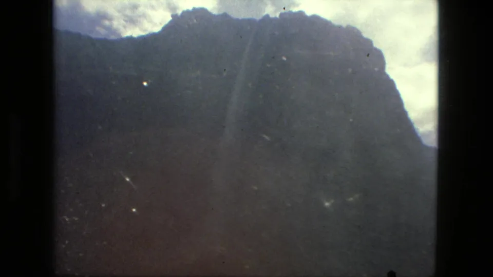 1979 8mm Brazil frame with a dark mountain face silhouetted against bright cloud cover and visible film specks.