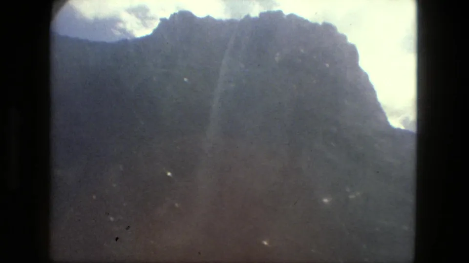 1979 8mm Brazil frame showing a broad mountain face beneath clouds, with a faint vertical streak near the center.