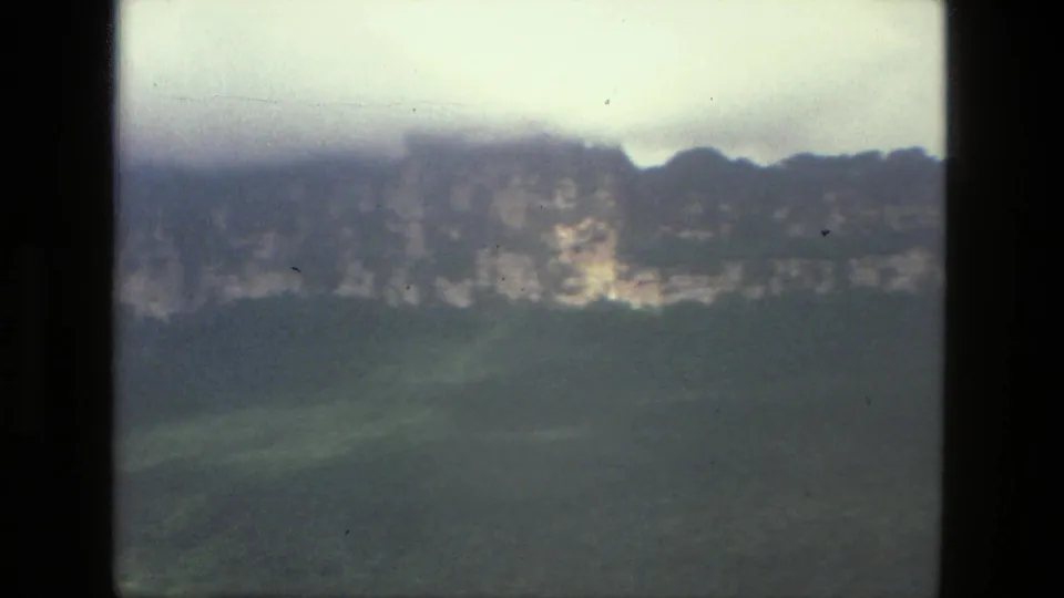 Soft 8mm frame from the 1979 Brazil landscape clip showing a distant ridge, pale rock bands, and green hillside below clouds.