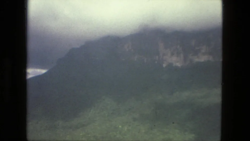 1979 8mm landscape frame showing a dark mountain shoulder, green lower slope, and low cloud cover in the supplied Brazil location.